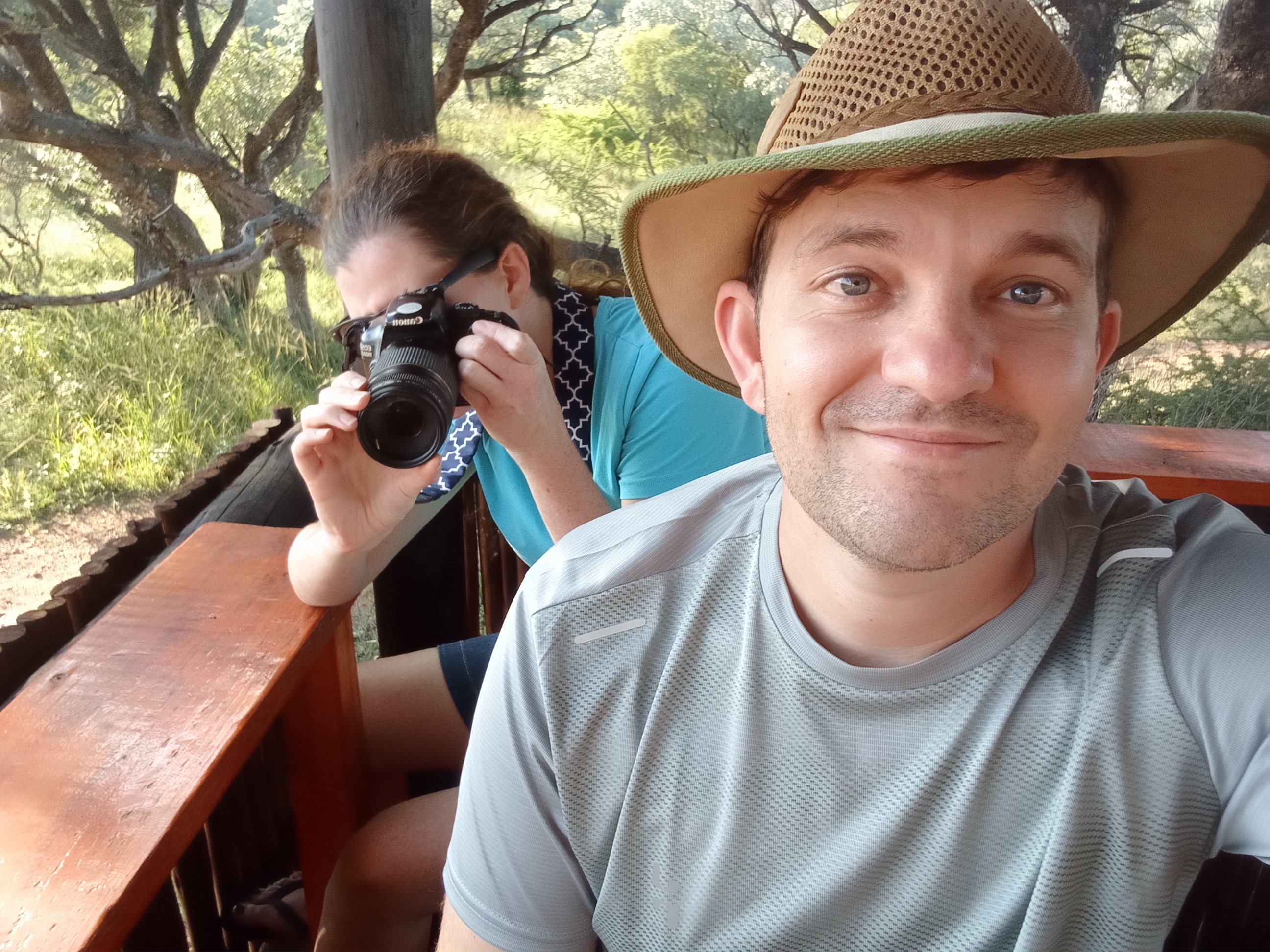 A man with hat looking at the camera smiling,
                        there is a woman with a camera behind him taking a photo